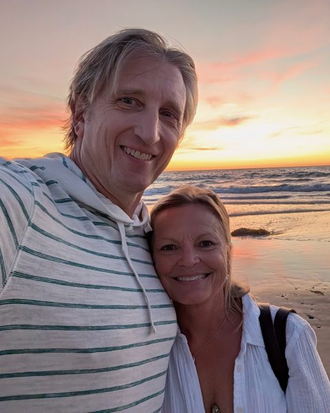 Nicky and Bob smiling in a selfie on the beach at sunset