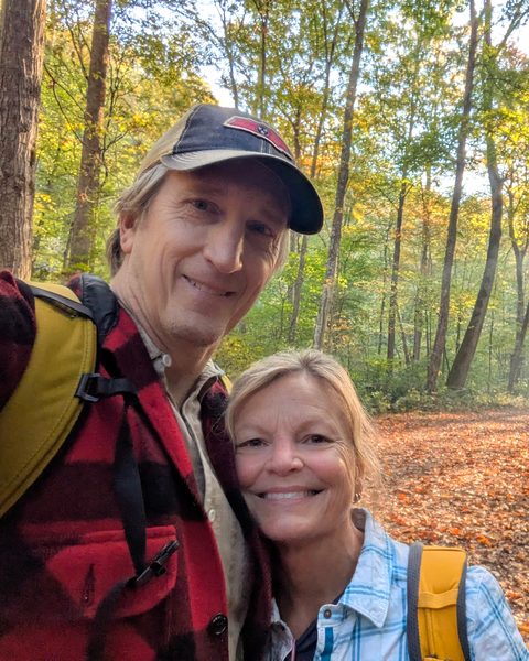 Nicky and Bob smiling in a selfie on a leaf-covered forest trail